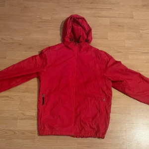 Röd vindjacka från Champion - Säljer en röd vindjacka från Champion i storlek S. Jackan har en huva och dragkedja framtill. Tillverkad av 100% nylon med foder av 100% polyester. Perfekt för blåsiga dagar.