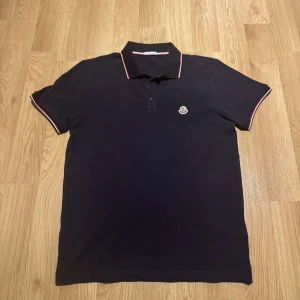 Moncler piké marinblå  - Snygg marinblå pikétröja från Moncler med klassisk krage och röd-vit randdetalj. Tröjan har korta ärmar och en liten Moncler-logga på bröstet. Perfekt för en stilren look. Storlek L men passar även M.