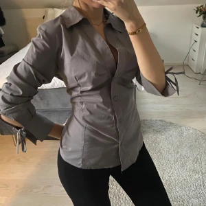 Grå blus med knytdetalj - Snygg grå blus med knytdetalj vid ärmsluten och en klassisk krage. Blusen har en figurnära passform och knappar framtill Uppskattar att den är i storlek S 