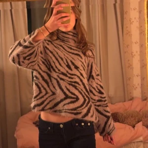 Zebramönstrad stickad tröja från H&M - Säljer en mysig zebramönstrad stickad tröja från H&M. Tröjan har en hög krage och långa ärmar, perfekt för kyligare dagar. Den är i en kombination av svart och lila/grå, vilket ger en trendig look. Passar bra till jeans eller kjol för en avslappnad stil. Jätte bra kvalitet
