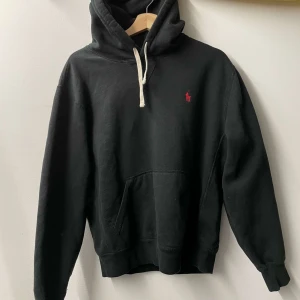 Svart hoodie från Ralph Lauren - Säljer en klassisk svart hoodie från Ralph Lauren med röd broderad logga på bröstet. Tröjan har en känguruficka och justerbar huva med vita snören. Perfekt för en stilren och bekväm look.