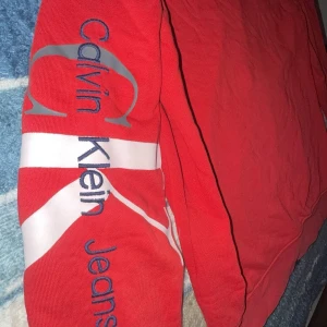 Röd hoodie från Calvin Klein Jeans - Säljer en snygg röd hoodie från Calvin Klein Jeans med dragkedja och tryck på ärmen. Perfekt för en casual look. Den har en klassisk passform och är tillverkad i mjukt material för extra komfort.