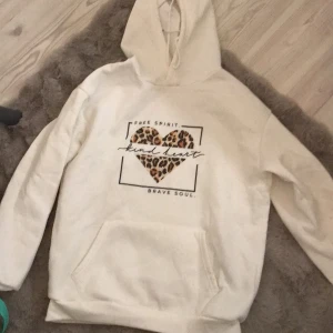 Vit hoodie med leopardhjärta - Vit hoodie med ett leopardmönstrat hjärta och texten 'Free Spirit, Kind Heart, Brave Soul’ på framsidan. Tröjan har en stor ficka framtill och en skön huva. Ganska nopprig annars ser den som ny ut. Om nån är intresserad tar jag bort det mesta av dem innan den skickas. 