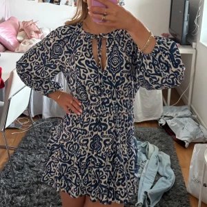 Klänning från Zara - Säljer en snygg kortklänning med blått och vitt mönster. Klänningen har långa ärmar och en volangdetalj vid fållen. Den har en knytning vid halsen. Storlek XS/S. Använd 2 gånger.