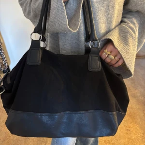 Svart axelväska med skinn detaljer - Snygg svart axelväska med skinn detaljer. Väskan har rejäla handtag och en stilren design som passar perfekt för vardagsbruk. Den är rymlig och praktisk för att bära med sig allt man behöver. Det sticker ut lite små trådar där uppe men det är inget man tänker på mycket