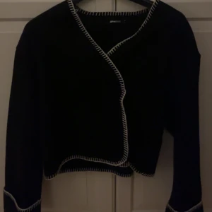 Svart kofta från Gina Tricot - Snygg svart kofta från Gina Tricot med vita kontrastsömmar. Den har en omlott-design och långa ärmar.