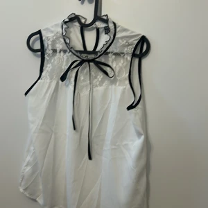 Vit ärmlös blus med spetsdetaljer - Säljer en söt vit ärmlös blus från Shein med spetsdetaljer och svart knytband vid halsen. Blusen har en elegant design med genombruten spets upptill och kontrasterande svarta kanter. Perfekt för en stilren look.