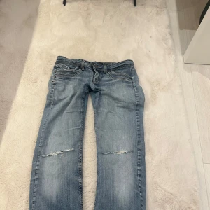 Blå jeans med slitningar - Snygga blå jeans med slitningar på knäna. De har en klassisk femficksdesign och en knappgylf. Perfekta för en avslappnad stil.💕