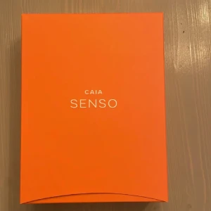 CAIA Senso Eau de Parfum - Säljer en elegant CAIA Senso Eau de Parfum i en fyrkantig orange flaska med matchande lock. Parfymen kommer i en stilren förpackning som också är orange. Perfekt för den som vill ha en doft med stil och karaktär.