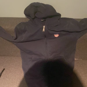 Mörkblå huvtröja från Gant - Säljer en mörkblå huvtröja från Gant med dragkedja och broderad logga på bröstet. Tröjan har en klassisk design med huva och långa ärmar. Perfekt för en avslappnad stil. 🧢