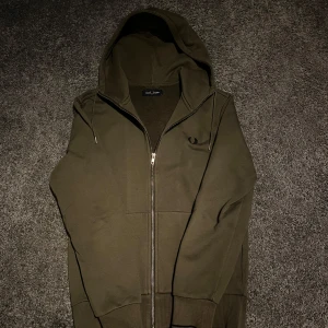 Olivgrön hoodie från Fred Perry - Säljer en snygg olivgrön hoodie från Fred Perry med dragkedja och huva. Perfekt för en avslappnad stil. Den har en diskret logga på bröstet och ribbade muddar vid ärmslut och nederkant. Tveka inte och höra av er vid funderingar! Priset är ej hugget i sten!🙌🏻