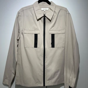 Beige jacka från studio total - Snygg beige jacka från Studio Total med dragkedja framtill och två bröstfickor med svarta detaljer. Jackan har en klassisk krage och långa ärmar, perfekt för en stilren look. Tunn och lämplig för vår. 