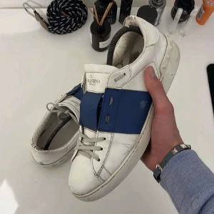 Valentino Open sneakers i vitt och blått - Säljer ett par snygga Valentino Open sneakers i vitt med en bred blå rem över snörningen. Skorna har en klassisk design med rund tå och är tillverkade i skinn. Perfekta för en stilren look.