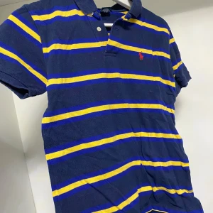 Blå och gul randig pikétröja från Ralph Lauren - Snygg blå pikétröja från Ralph Lauren med gula ränder och klassisk krage. Tröjan har korta ärmar och en liten röd logga på bröstet. Perfekt för en avslappnad stil.