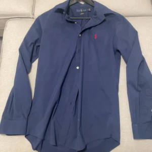 Ralph Lauren skjorta  - Snygg blå skjorta från Ralph Lauren i slim fit. Skjortan har långa ärmar och en klassisk krage. Den är dekorerad med en röd broderad logotyp på bröstet. Perfekt för en stilren look.