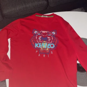 Röd tröja från Kenzo med tigerbroderi - Säljer en snygg röd tröja från Kenzo med ett coolt tigerbroderi på framsidan. Tröjan har långa ärmar och en rund halsringning. Perfekt för att ge din outfit en färgklick!