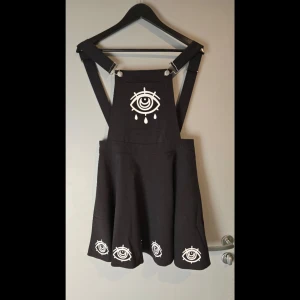 Svart klänning med ögonmotiv - Snygg svart klänning med unikt ögonmotiv i vitt. Klänningen har justerbara axelband och en skaterkjol som ger en cool look. Perfekt för dig som vill sticka ut med en edgy stil.