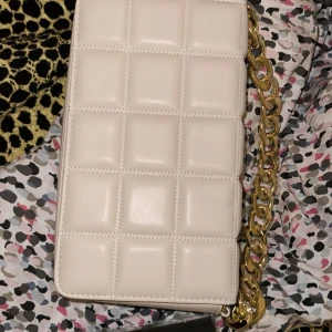 Beige axelväska med kedja - Snygg vit axelväska med quiltat mönster och en elegant guldkedja som axelrem. Perfekt för att ge en stilren touch till din outfit. Väskan har en praktisk dragkedja för säker förvaring av dina saker.