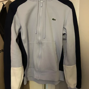 Ljusblå hoodie från Lacoste - Säljer en stilren ljusblå hoodie från Lacoste med dragkedja och mörkblå detaljer på ärmarna. Tröjan har en klassisk krokodil-logga på bröstet och vita muddar vid ärmsluten. Perfekt för en avslappnad stil. Kvitto finns från Lacoste 
