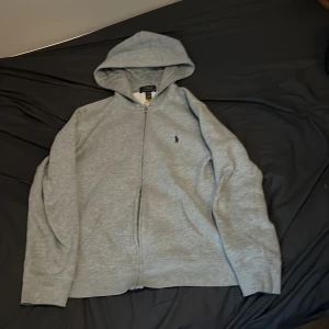 Grå hoodie från Ralph Lauren - Säljer en stilren grå hoodie från Ralph Lauren med dragkedja och den klassiska loggan broderad på bröstet. Perfekt för en avslappnad look. Hoodien har långa ärmar och en bekväm passform. Nästan oanvänd. Köpt från kids brand store. Modellen är cirka 171 cm lång.