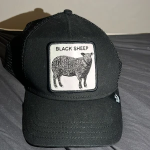 Svart keps från The Farm - Snygg svart keps från The Farm med ett coolt 'Black Sheep'-märke framtill. Kepsen har ett nätmaterial baktill för extra ventilation och justerbar passform. Perfekt för en avslappnad stil.