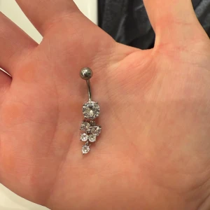 Silverfärgad navelpiercing med glittriga stenar - Elegant navelpiercing i silverfärg med flera glittriga stenar i olika storlekar. Helt oanvänd, säljes då den inte kommer till andvänding.