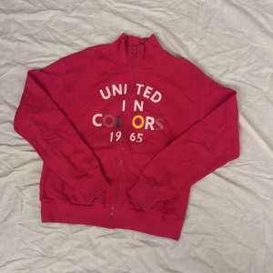 Röd hoodie med dragkedja från United Colors - Säljer en snygg röd hoodie med dragkedja från United Colors. Den har texten 'United in Colors 1965' tryckt på framsidan. Tröjan har långa ärmar och praktiska fickor framtill. Perfekt för en avslappnad stil.