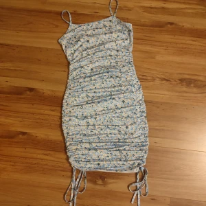 Blommig klänning från Shein - Säljer en söt blommig klänning från Shein i ljusblått med små gula blommor. Klänningen har smala axelband och en åtsittande passform med knytband i nederkant. Längden på klänningen går att justera med hjälp av banden vilket gör att man kan ha den som en kort klänning eller ända ner till under kväna. Perfekt för sommarens varma dagar! 🌼