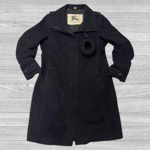 Svart kappa från Burberry - Elegant svart kappa från Burberry med klassisk design. Kappan har långa ärmar och ett medföljande bälte för en stilren look. Perfekt för kyligare dagar med sin tidlösa stil och högkvalitativa material.