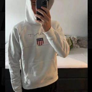 Vit hoodie från GANT - Säljer en stilren vit hoodie från GANT med deras logotyp och ett emblem på bröstet. 