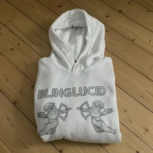 Vit hoodie från Blinglucid - Säljer en vit hoodie från Blinglucid med glittrig text och två änglar på framsidan. Den har en klassisk känguruficka och en bekväm huva. Perfekt för en stilren och avslappnad look. Det är en liten smink fläck på insidan utav luvan men de är inget man ser när ttröjan är på eller när luvan är på!ny pris 700!
