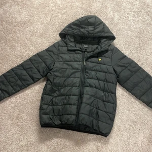 Svart pufferjacka från Lyle & Scott - Säljer en svart pufferjacka från Lyle & Scott Junior. Jackan har en quiltad design med dragkedja och huva. Perfekt för kyligare dagar. Är dock lite trasig i armhålorna så priset kan diskuteras 