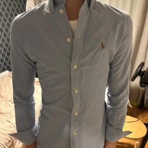 Ralph lauren Ljusblå skjorta  - Snygg ljusblå skjorta från Ralph Lauren med en broderad logga på bröstet. Skjortan har långa ärmar och knappar framtill. Perfekt för en stilren look. Den är storleken S och slim fit, passar xs-s