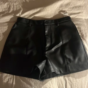 Svart skinnshorts  - Snygg svarta shorts i skinn med hög midja och stilren design. Shortsen har en knapp och dragkedja framtill samt hällor för bälte. Perfekt för en trendig look!