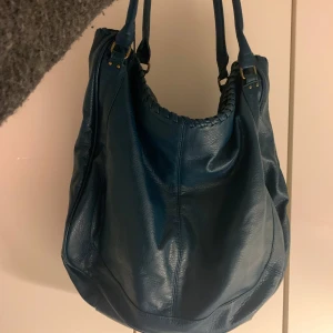 Blå axelväska från H&M - Snygg blå axelväska från H&M. Väskan har flätade detaljer vid öppningen. En innerficka med blixtkejda och två innerfack