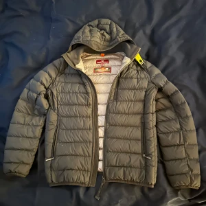 Svart dunjacka från Parajumpers - Säljer en svart superlätt dunjacka från Parajumpers med huva och dragkedja. Jackan har en cool patch på ärmen och är perfekt för kyliga dagar. Den är designad för att vara både stilren och funktionell.