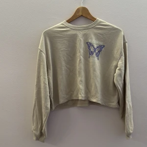 Beige croppad tröja med fjärilstryck - Säljer en beige croppad tröja med ett coolt fjärilstryck och texten 'Stay Magical' på framsidan. Tröjan har långa ärmar och en avslappnad passform. Perfekt för en chill dag eller när du vill känna dig magisk 🦋.Ungdoms storlek 158-164