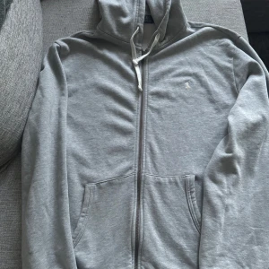 Grå hoodie från Polo Ralph Lauren - Säljer en stilren grå hoodie från Polo Ralph Lauren med dragkedja och broderad logga på bröstet. Tröjan har en klassisk design med huva och fickor framtill. Perfekt för en avslappnad look.