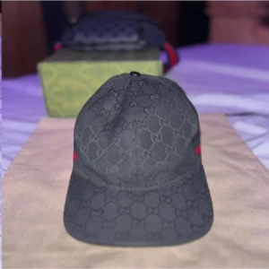 Svart keps från Gucci - Snygg svart keps från Gucci med det ikoniska GG-mönstret. Kepsen har en justerbar rem baktill och detaljer i rött och grönt på sidorna. Perfekt för att ge en lyxig touch till din outfit. Väska medföljer ej!