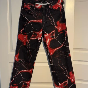 Jaded London Lightning Jeans - Unika svarta byxor från Jaded London med ett  rött och vitt blixt mönster. Tillverkade i 100% bomull för en bekväm passform. Perfekta för dig som vill sticka ut med en cool stil.