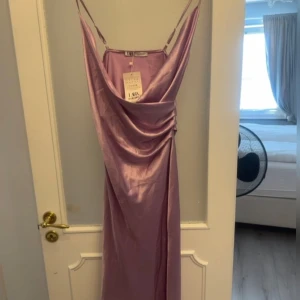 Ljuslila långklänning från LARA - Elegant ljuslila långklänning från LARA i ett glansigt material. Klänningen har smala axelband och en draperad framsida som ger en lyxig känsla. Perfekt för speciella tillfällen.