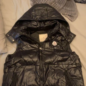 Svart dunväst från Moncler - Snygg svart dunväst från Moncler med huva och dragkedja. Västen har en quiltad design och en broderad logotyp på bröstet. Perfekt för kyligare dagar.