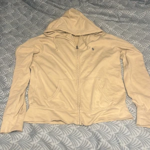 Beige huvtröja från Ralph Lauren - Säljer en snygg beige zipp från Ralph Lauren med dragkedja och broderad logga på bröstet. Tröjan har långa ärmar och en praktisk huva med dragsko. Perfekt för en avslappnad stil. 