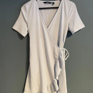 Ljus omlottklänning från Vero Moda - Säljer en stilren ljus omlottklänning från Vero Moda. Klänningen har korta ärmar och en v-ringad design med volangdetaljer längs kanten. Perfekt för en somrig look! 💫