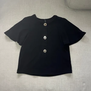 Svart kortärmad blus med knappar - Snygg svart blus med fladdrigare armar och stora dekorativa knappar framtill. Perfekt för en stilren look. Mycket gott skick!