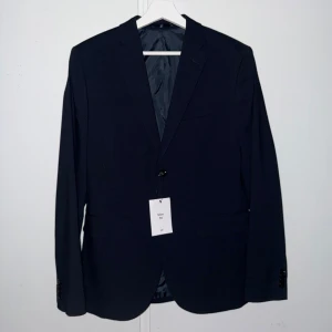 Mörkblå kavaj - Elegant mörkblå kavaj med klassisk design från hm. Perfekt för formella tillfällen. Kavajen har en enkelknäppt stil med två knappar framtill och fickor på sidorna. Passar utmärkt för en stilren look. Storlek 46. Den är helt ny och aldrig använd. Köpt för 499kr. 