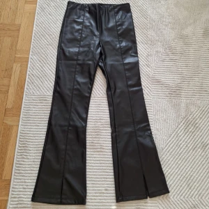 Svarta skinnbyxor med slits - Snygga svarta skinnbyxor med hög midja och bootcut-stil. Byxorna har en stilren design med slitsar vid bensluten som ger en trendig touch. Perfekta för att höja din outfit till nästa nivå.