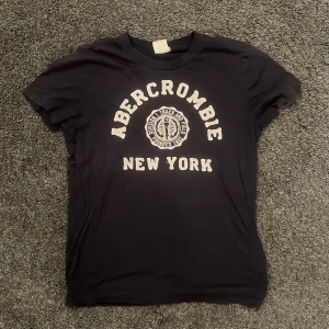 Svart t-shirt från Abercrombie & Fitch - Snygg svart t-shirt från Abercrombie & Fitch med tryck 'Abercrombie New York' i vitt. Perfekt för en avslappnad stil. T-shirten har en normal passform och är kortärmad.