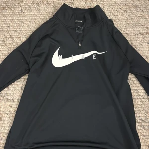 Svart Nike Dri-FIT tröja - Säljer en svart Nike Dri-FIT tröja med halv dragkedja och stort Nike-logotyp på framsidan. Perfekt för löpning och träning med långärmad design och bekvämt material.