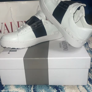 Valentino Garavani sneakers - Snygga Valentino Garavani sneakers i vitt med en bred svart rem över ovansidan. Skorna har en stilren design med snörning och subtila detaljer på sulan. Perfekta för en trendig look.